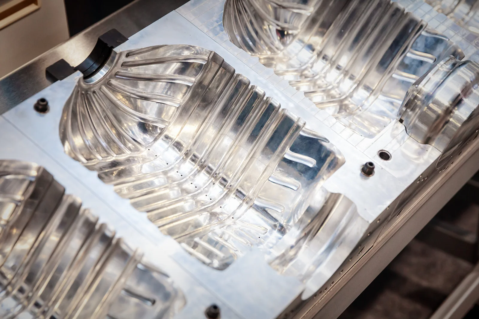 Three translucent molded plastic containers are shown in a tray fixture under bright lighting, suggesting mass production in an injection molding setup; the image highlights molding as a manufacturing process prone to defects such as porosity or leakage. The containers appear uniform in shape, with fine seams and surface details visible. The mold or fixture holding them conveys the industrial context. This visual supports discussion of why molded parts require rigorous leak testing in common manufacturing processes to ensure airtight or watertight integrity.